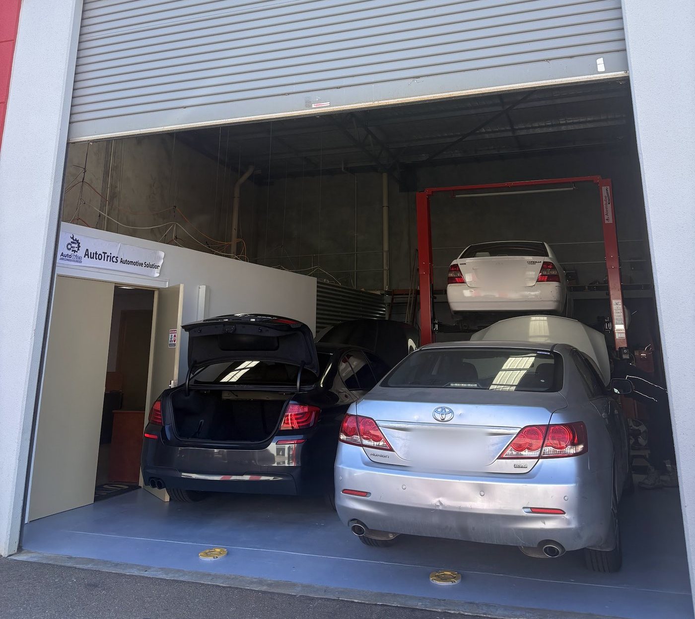 Inside the AutoTrics bay: a car raised on the red two-post lift and two cars in for work, one with its boot open