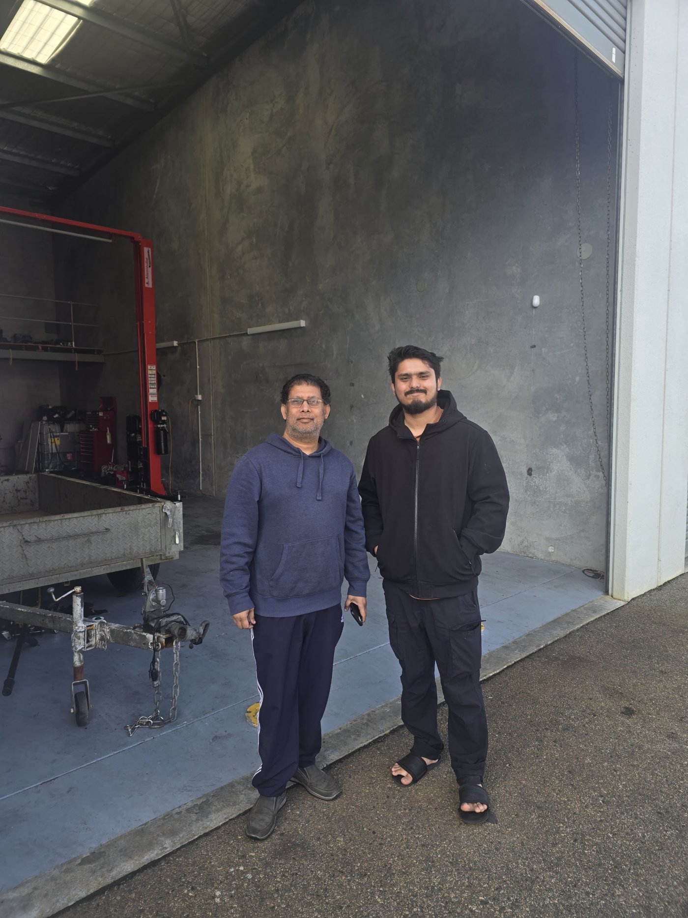 Two members of the AutoTrics team standing at the open roller door of the workshop, the red two-post lift behind them